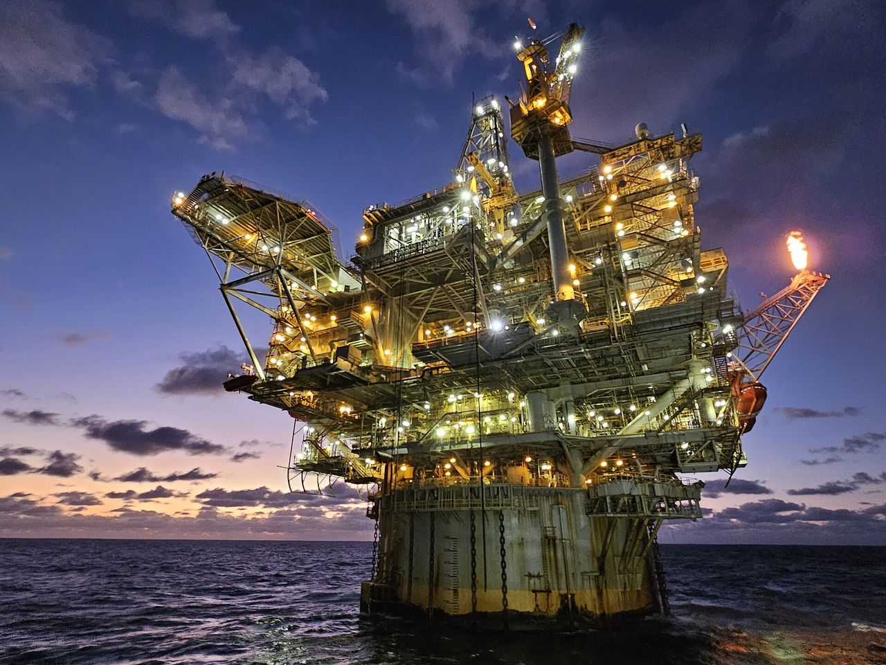 An offshore oil and gas platform lit up at dusk, with a gas flare against a purple evening sky